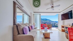 A modern living room with a sofa, coffee table, and red chair. A ceiling fan and TV are visible. - The Park 506 | 2BR/2BA | Puerto Vallarta | Pool, Gym (Puerto Vallarta)