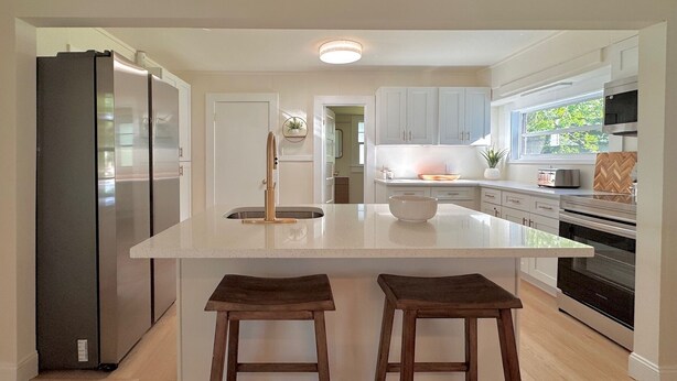 A modern kitchen with a large island, wooden stools, stainless steel appliances, and a view of the outdoors. - Blue Cottage On Parker- by CasaDuran (Lakeland)