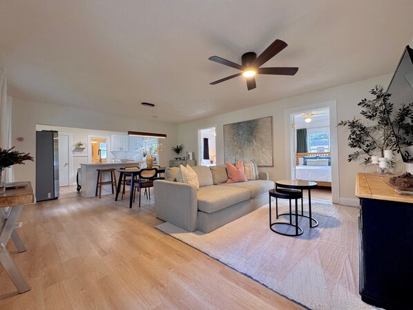 A modern living room with a grey sofa, a dining area with a table and chairs, a kitchen in the background, and a ceiling fan.