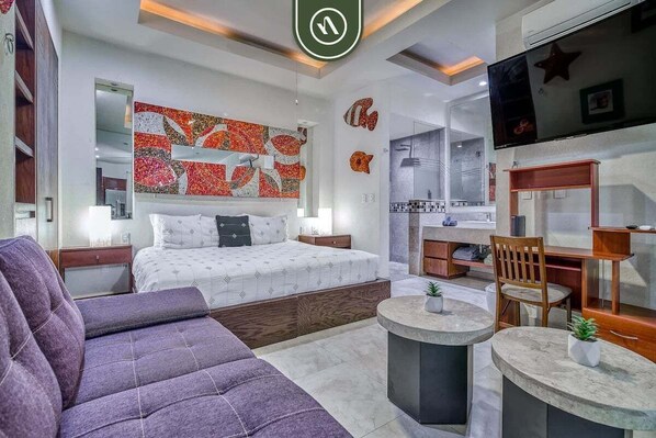 A modern hotel room with a bed, a sofa, a dining area, and a TV.