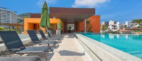 Un espace aménagé au bord de la piscine, avec des chaises longues et des parasols, une piscine à l’eau d’un bleu limpide et un bâtiment moderne arborant un logo distinctif.