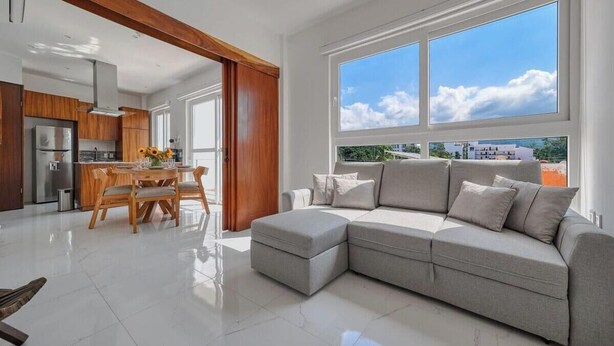 A modern living room with a grey sectional sofa, a dining area with wooden furniture, and large windows offering a view of the city. - Amazing 1BR - Rooftop Pool in Versalles (Puerto Vallarta)