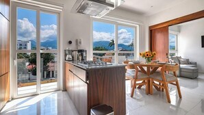 A modern kitchen with large windows, a dining area with wooden chairs, and a view of mountains. - Amazing 1BR - Rooftop Pool in Versalles (Puerto Vallarta)