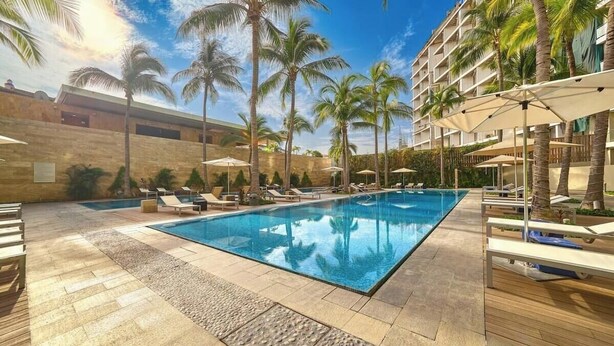 A rectangular swimming pool with lounge chairs and umbrellas, surrounded by palm trees and a stone wall. - Luxury 2BR Condo in Marina - Pool - Zen Rooftop (Puerto Vallarta)