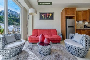 Ein Wohnzimmer mit einem roten Sofa, zwei Korbstühlen, einem Couchtisch mit Vasen, einer Küche mit Holzschränken und Blick auf die Stadt durch große Fenster.