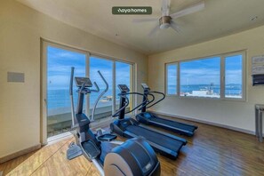 Ein Fitnessraum mit Laufbändern und Crosstrainern, großen Fenstern mit Meerblick und einem Deckenventilator.