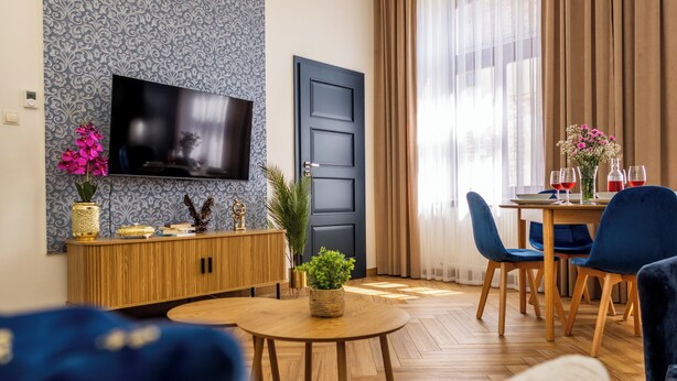 Una sala moderna con televisor de pantalla plana, un mueble de madera, una zona de comedor con sillas azules y un ventanal con cortinas.