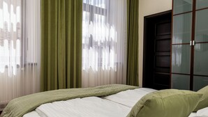 Una cama bien hecha con una colcha verde y almohadas, una ventana con cortinas translúcidas y un armario de madera oscura.