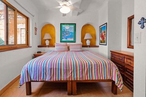 Quarto com cama grande, duas luminárias de cabeceira, ventoinha no teto e janela com vista para uma parede de tijolos.
