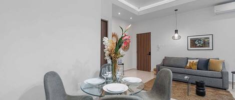A modern living room with a grey sofa, a glass dining table with four chairs, and a vase of flowers.