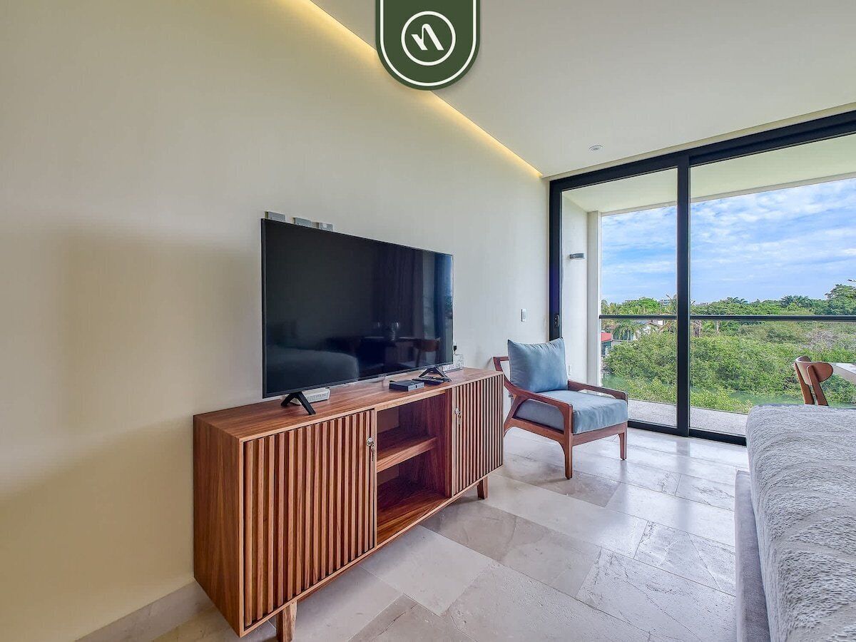 Een moderne woonkamer met een flatscreen televisie op een houten console, een comfortabele fauteuil en een groot raam met uitzicht op groen.