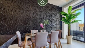 A modern dining area with a geometric wall design, a large potted plant, and a glass table with chairs. - Amazing 1 BR Condo with Terrace - Rooftop Pool (Puerto Vallarta)
