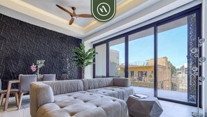 A modern living room with a large sectional sofa, a dining area with a table and chairs, and a large window offering a city view. - Amazing 1 BR Condo with Terrace - Rooftop Pool (Puerto Vallarta)