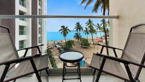 Un balcon avec deux chaises et une petite table, donnant sur une plage bordée de palmiers et surplombant un ciel bleu azur.