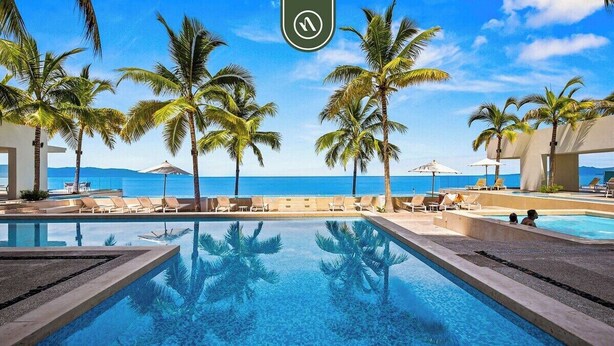 Une vue sur la piscine, avec des palmiers, des chaises longues et une vue dégagée sur l’océan.