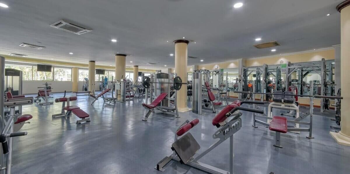 A spacious gym with various exercise machines, large windows, and a polished floor.