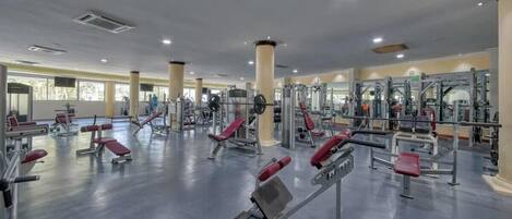 A spacious gym with various exercise machines, large windows, and a polished floor.