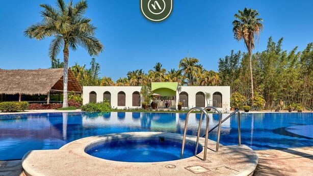 Una piscina de borde circular, palmeras y un edificio con ventanas arqueadas al fondo.