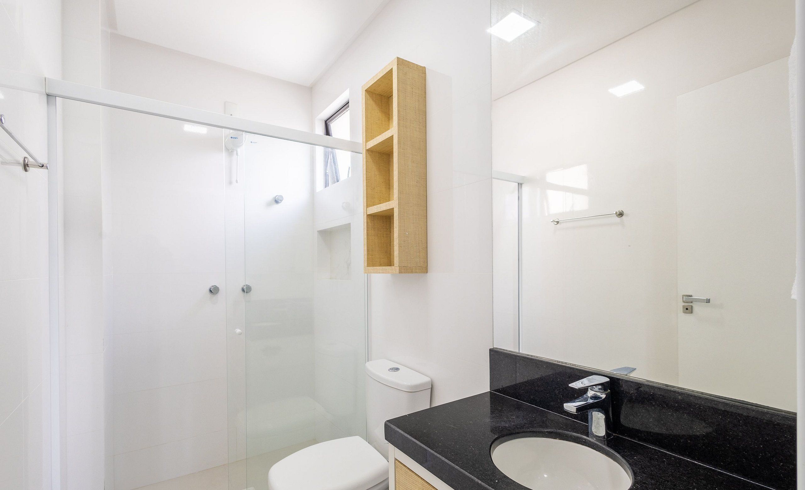 A modern bathroom with a glass shower enclosure, a white toilet, a black countertop with a sink, and a wooden shelf above the toilet.