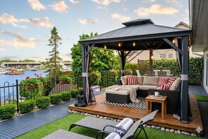 A covered outdoor seating area with a view of a body of water, a dock, and a row of houses. - Private Waterfront Paradise- Pool, Sauna, Hot Tub, Pontoon Boat, Billiard, Bikes (Beaverton)