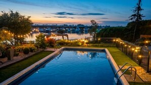 Une piscine avec vue sur un port de plaisance et des bateaux, entourée d’une pelouse bien entretenue et d’un éclairage décoratif.