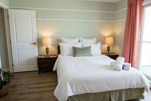 Cama bem arrumada com travesseiros, duas luminárias de cabeceira, mesa de cabeceira e janela com cortinas.