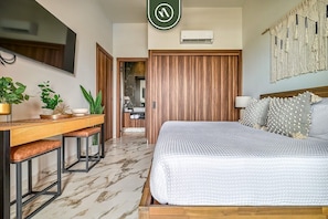 A bedroom with a bed, a desk with chairs, a TV, and an air conditioning unit.