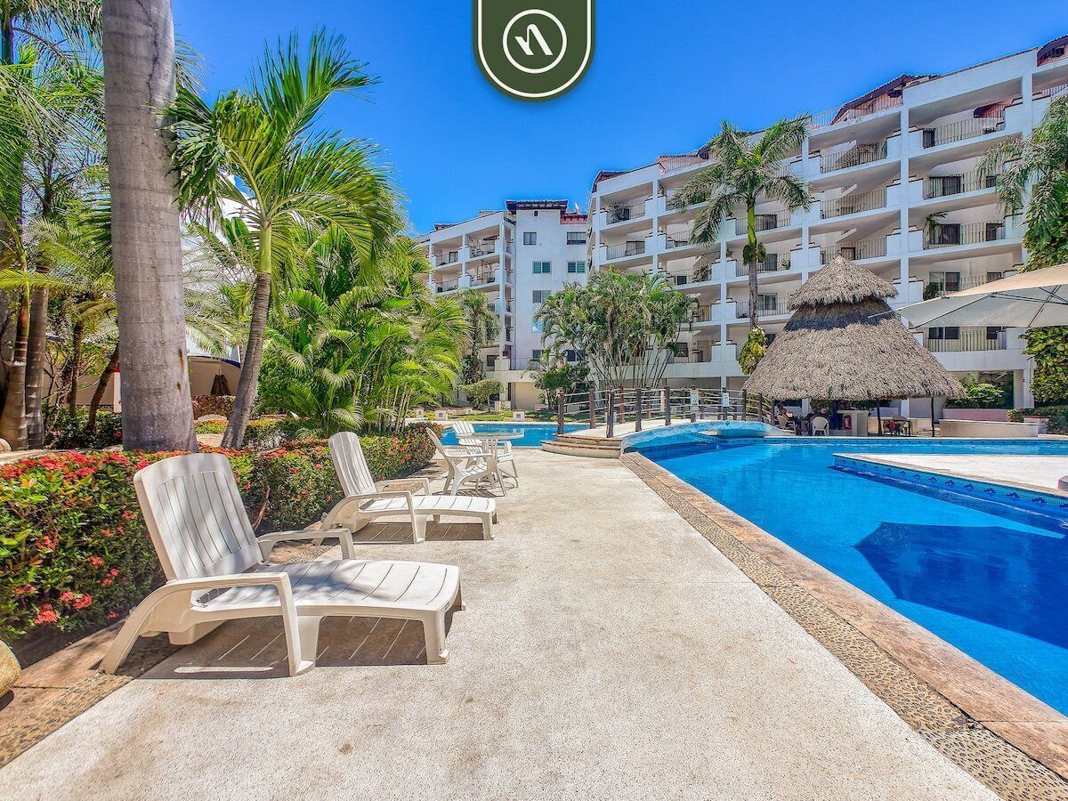 A resort pool area with lounge chairs, a thatched-roof structure, and a multi-story building with balconies.