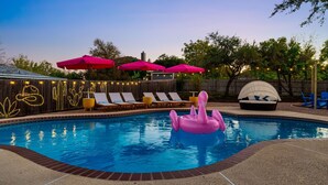 Una piscina en el patio trasero con sillas de descanso, un flotador con forma de flamenco rosa y luces colgantes.