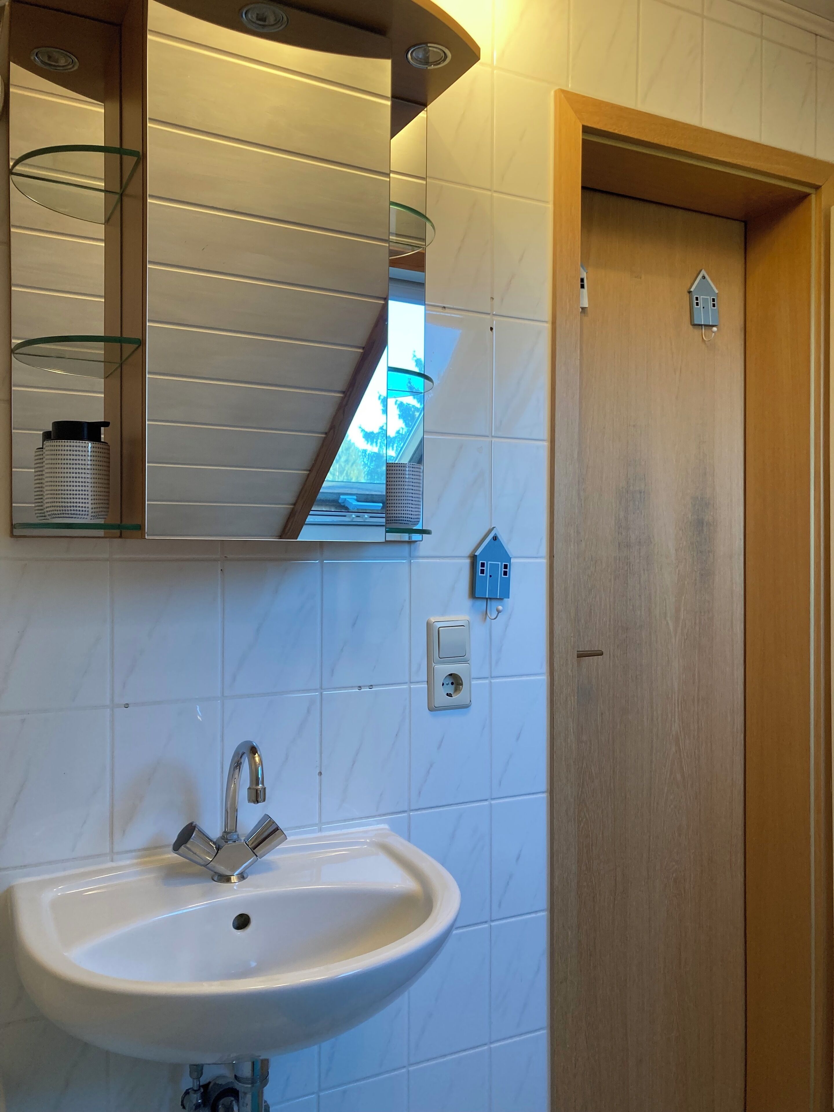 Une salle de bain avec un lavabo blanc, un miroir doté d’étagères et une porte en bois.