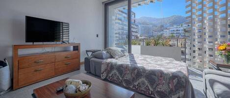 A modern hotel room with a large bed, a TV on a wooden dresser, a balcony with a city view, and a dining table with a bowl of fruit.