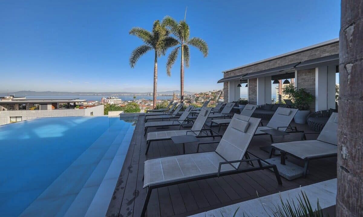 Une piscine sur le toit, agrémentée de chaises longues et de palmiers, offrant une vue panoramique sur la ville.