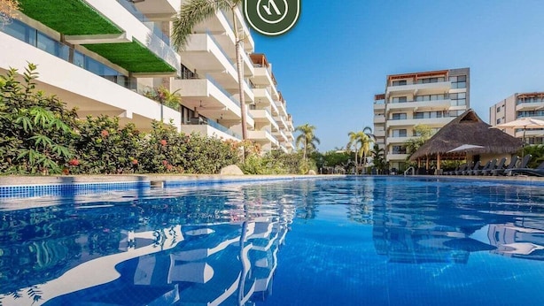 Una piscina con superficie de agua azul cristalina, rodeada de edificios modernos con balcones y vegetación.