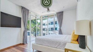 A bedroom with a bed, a bedside table with a lamp, a TV mounted on the wall, and a view of a building through a large window. - The Ava Unit 16 | 2BR/3BA Puerto Vallarta, pool, gym, hot tub (Puerto Vallarta)