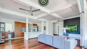 Uma sala de estar ampla, com um sofá cinzento grande, piso de madeira, ventiladores de teto e uma televisão.