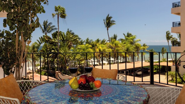 A table with a fruit plate, two wine glasses, and a view of the beach and ocean. - 303-VistadelSol | 2BR/2BA Puerto Vallarta, Ocean View (Puerto Vallarta)