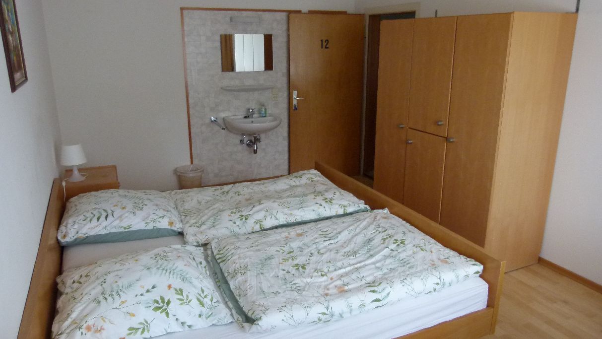 Ein Zimmer mit Doppelbett, einem Holzschrank, einem kleinen Nachttisch mit Lampe und einem fest installierten Waschbecken.