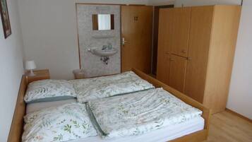 Ein Zimmer mit Doppelbett, einem Holzschrank, einem kleinen Nachttisch mit Lampe und einem fest installierten Waschbecken.