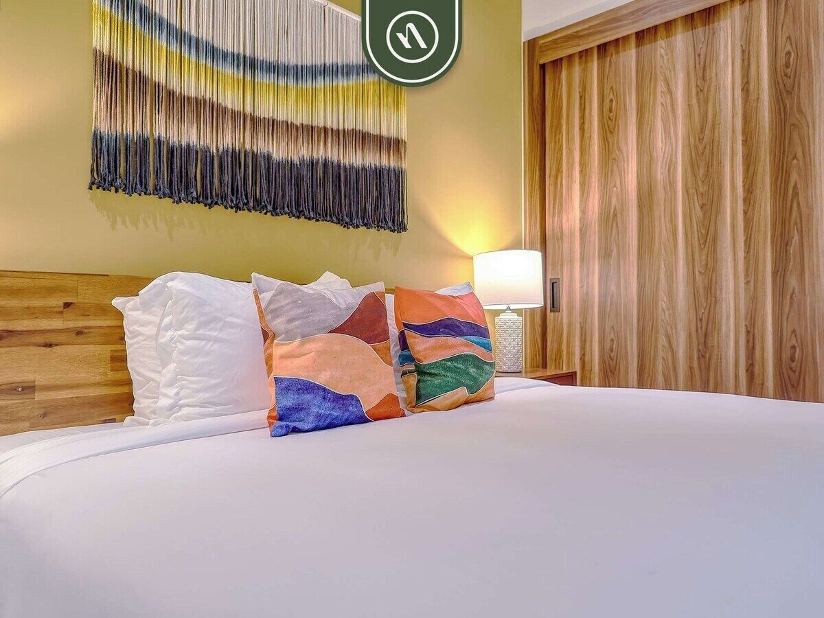 Une chambre d’hôtel avec une tête de lit en bois, un lit avec une literie blanche, des coussins colorés, une œuvre d’art abstraite accrochée au mur et une armoire en bois.