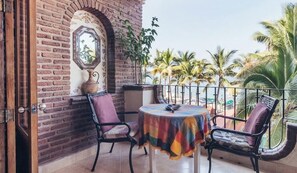 A balcony with a brick wall, a round table with a colorful tablecloth, two chairs, and a view of palm trees and the sea.