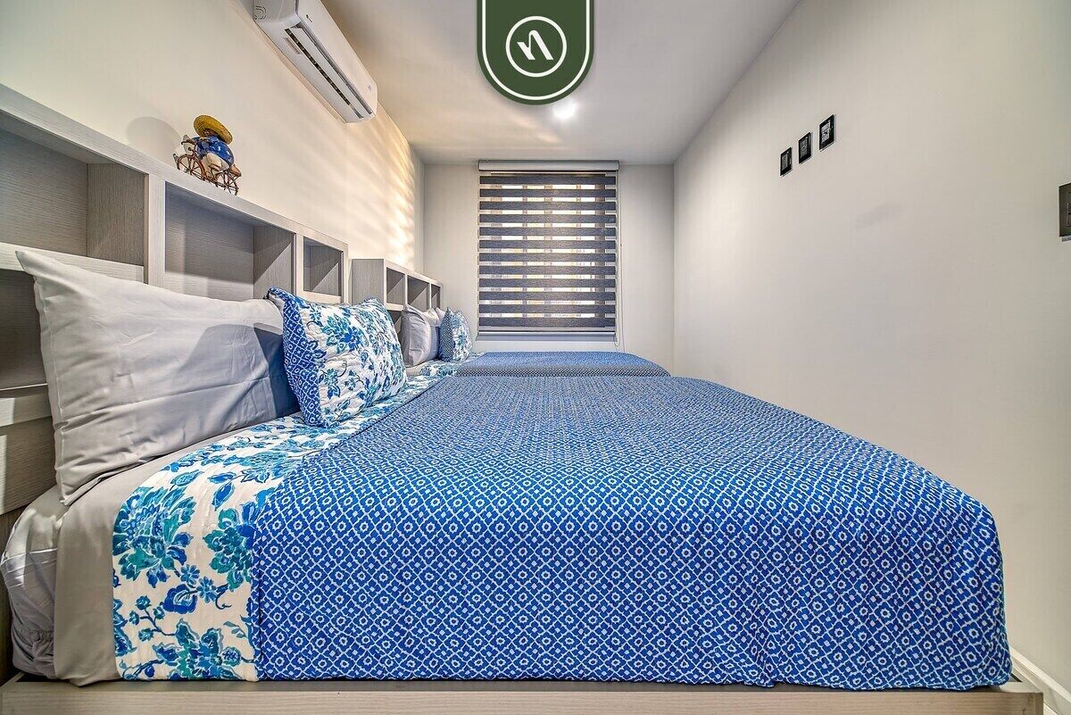 A bedroom with a bed, white walls, a window with blinds, and a small shelf with decorative items.