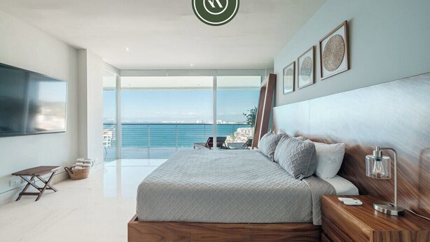 A modern bedroom with a large bed, a wooden nightstand, and a view of the sea. - Paramount Bay PH 5 | 3BR/4BA Penthouse, PV | Ocean Views, Rooftop Pool (Puerto Vallarta)