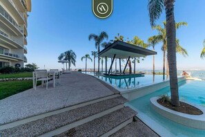 A resort pool area with a covered seating area, palm trees, and a view of the ocean. - Paramount Bay PH 5 | 3BR/4BA Penthouse, PV | Ocean Views, Rooftop Pool (Puerto Vallarta)