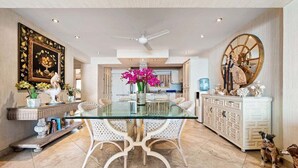 A spacious dining area with a glass table and white chairs, a ceiling fan, and a decorative wall piece.