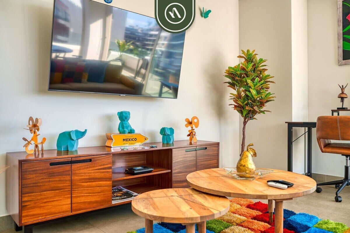 A modern living room with a TV mounted on the wall, a wooden cabinet with decorative items, a round wooden table, and a colorful rug.