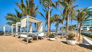 Un hotel a la vora de la platja, amb gandules blanques, palmeres i una vista despejada de l'oceà.