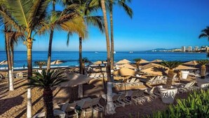Une plage avec des chaises longues et des parasols, des palmiers, et une vue sur la mer et les bâtiments qui se dressent au loin.