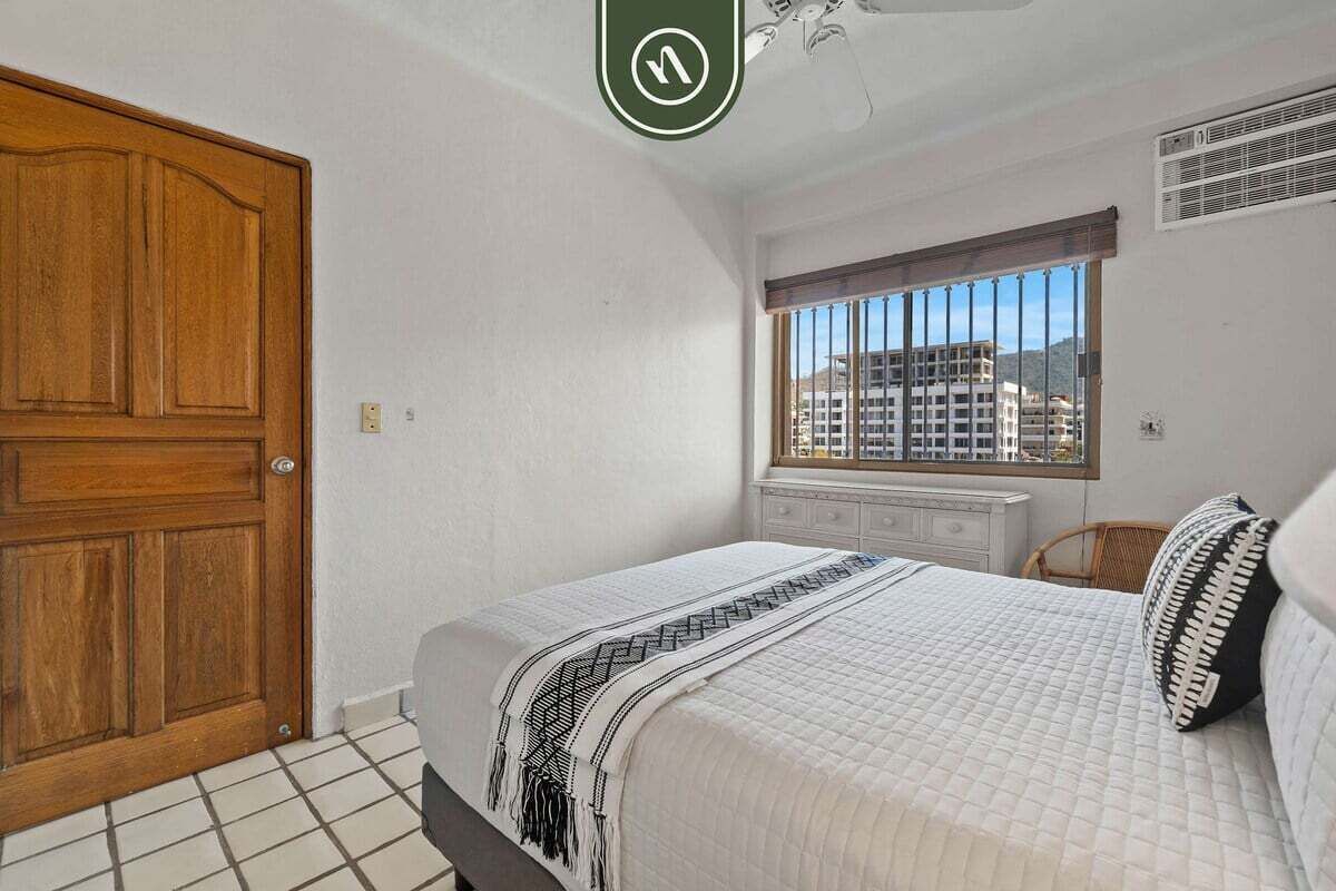 A bedroom with a large wooden door, a window with a view of buildings, a bed with a patterned blanket, and a white dresser.
