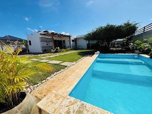 Una piscina rectangular con borde de piedra, rodeada por un césped cuidado y un pequeño edificio de fachada blanca.