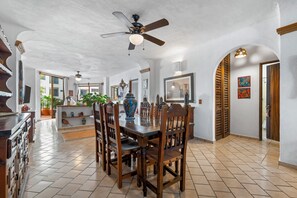 A spacious dining area with a high ceiling fan, wooden chairs, a large dining table, and a view of the outside through an open doorway.